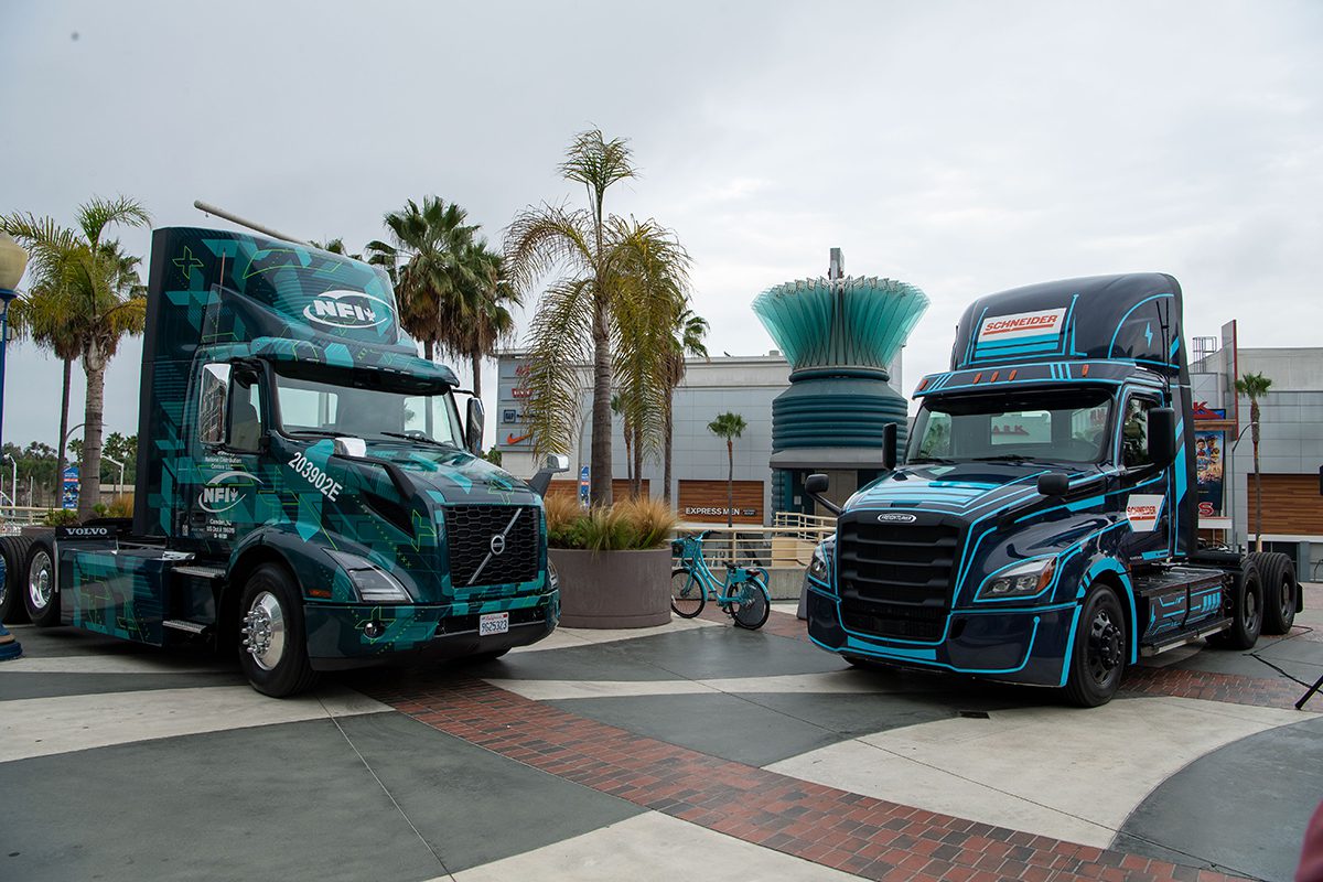 Volvo and Freightliner battery-electric semi trucks parked outside on concourse